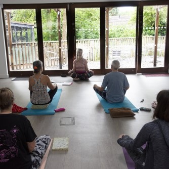 Five cross legged people sit on yoga mats beside a large window overlooking the park